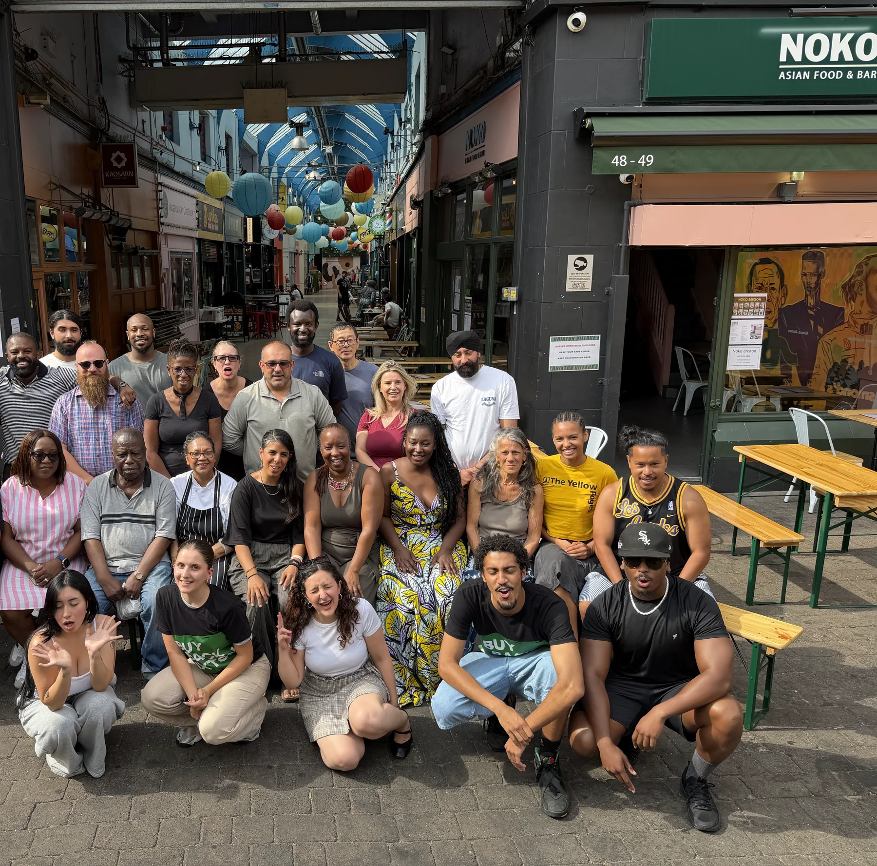 Around thirty traders and campaigners posed together in the entrance to Brixton Village, paper lanterns strung down the arcade behind them, market units either side.