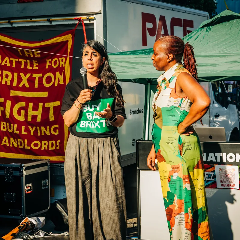 A speaker on stage at the rally in a brightly patterned dress, beside a banner reading The Battle for Brixton: Fight Bullying Landlords.