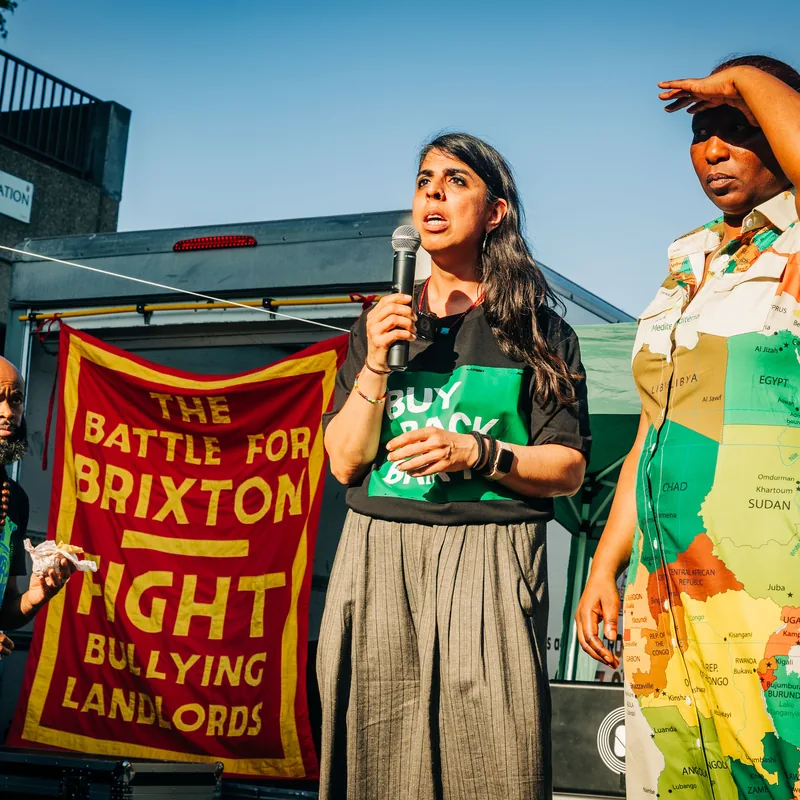 Two campaigners on stage, one speaking into a microphone, in front of the Battle for Brixton banner.