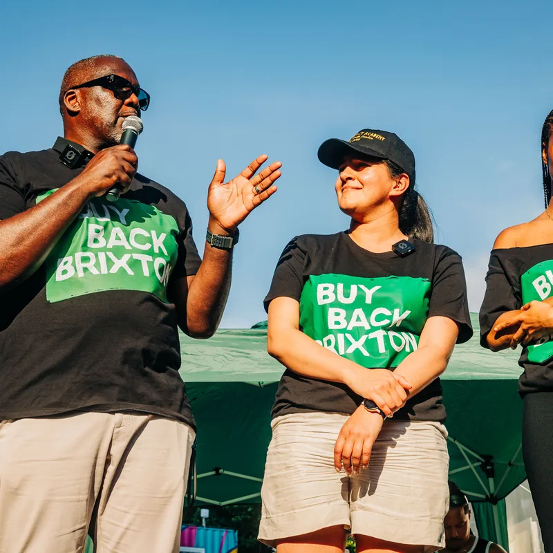 A campaigner speaking into a microphone on stage, flanked by two others in Buy Back Brixton T-shirts.