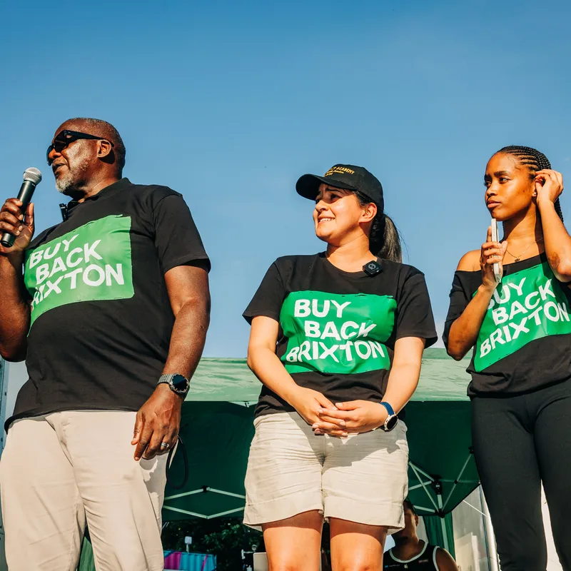 Campaigners standing together on stage in Buy Back Brixton T-shirts under a bright blue sky.