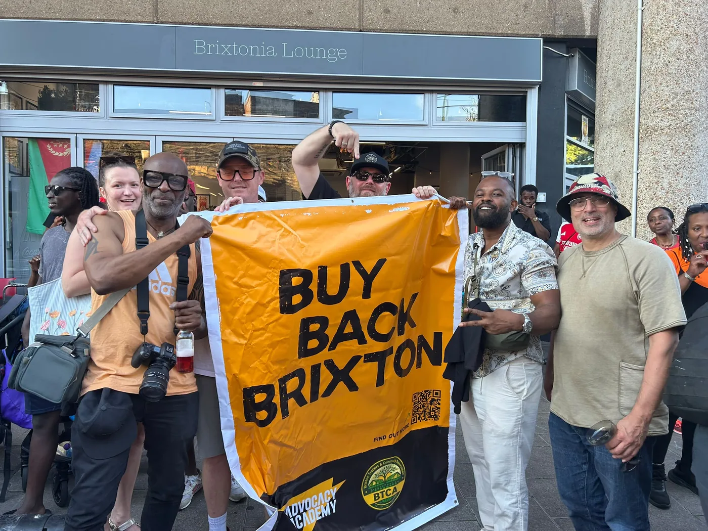 Supporters and traders gathered on Brixton Station Road behind a large orange Buy Back Brixton banner, smiling and posing for the camera.