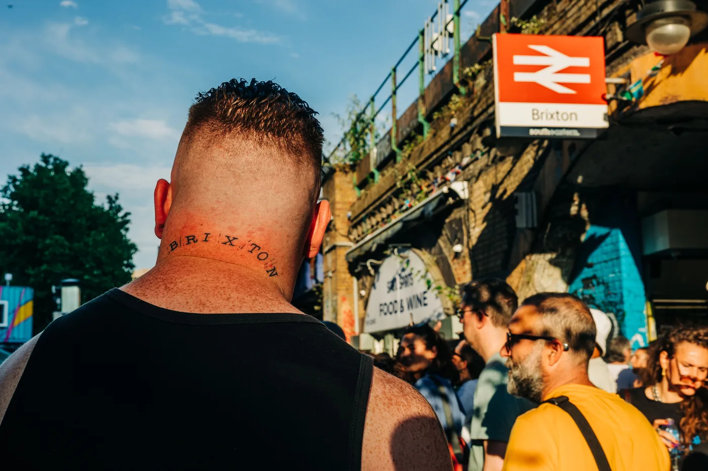 A man at the rally with BRIXTON tattooed across the back of his neck, the Brixton station sign and the crowd ahead of him.